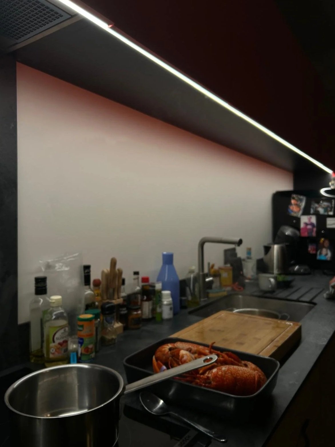 Une cuisine moderne avec un plan de travail noir, un évier, une planche à découper, des assiettes de crustacés, bouteilles et bocaux d'épices, une cafetière, un réfrigérateur avec des photos et des magnets.