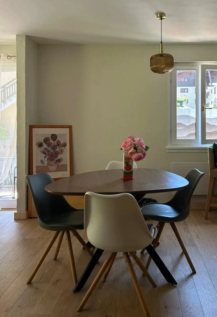 Salon avec une table en bois ronde, quatre chaises, une fenêtre, un vase avec des roses, un cadre d'œuvre d'art, et un luminaire suspendu.