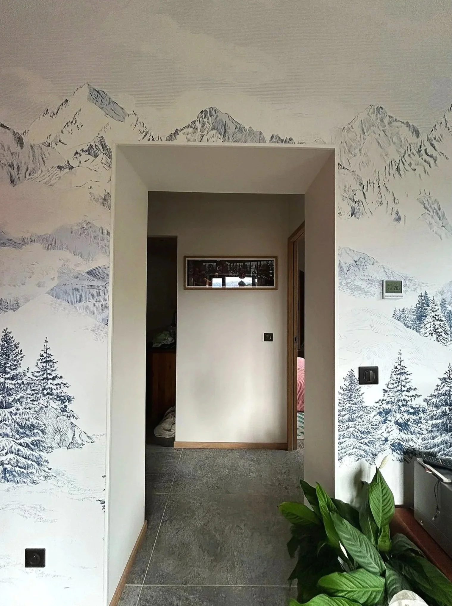 Une pièce avec un mur décoré d'une fresque représentant des montagnes enneigées et des arbres. Au centre, un mur blanc avec un miroir rectangulaire. À droite, une plante verte.