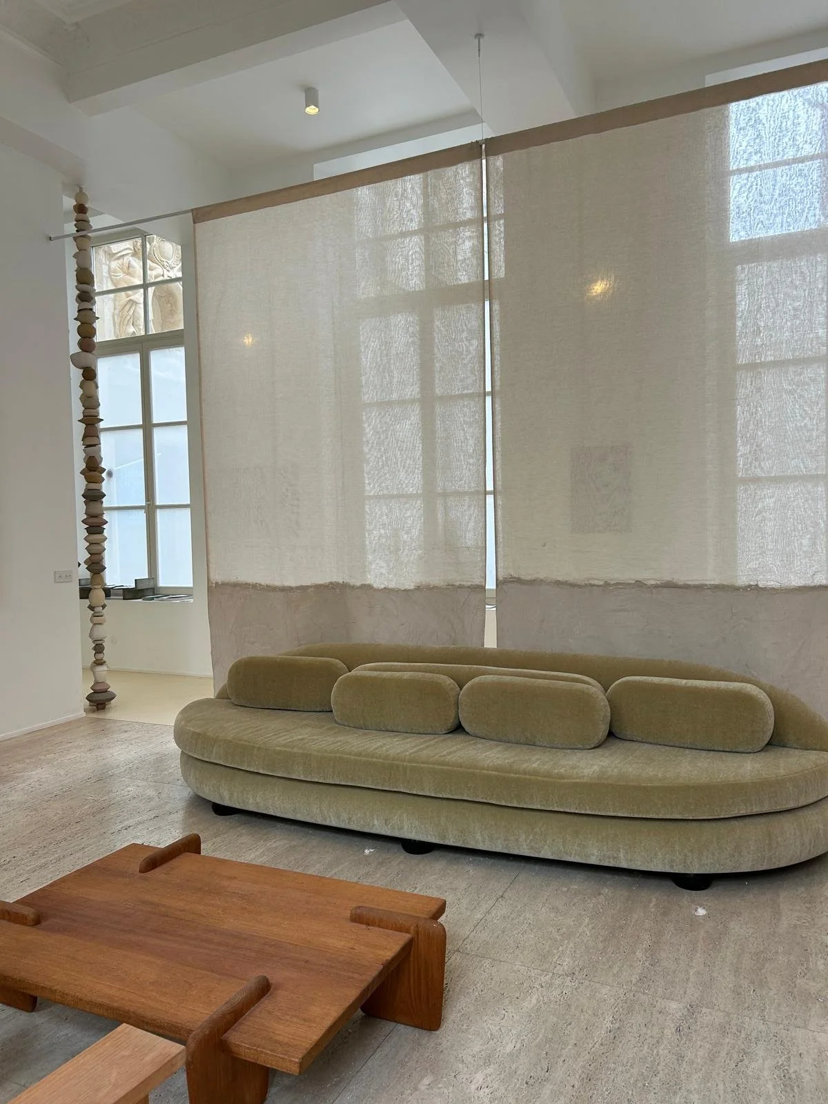 Intérieur moderne avec un divan beige à forme ovale, séparé par une grande tenture beige transparente, face à une fenêtre laissant passer la lumière.