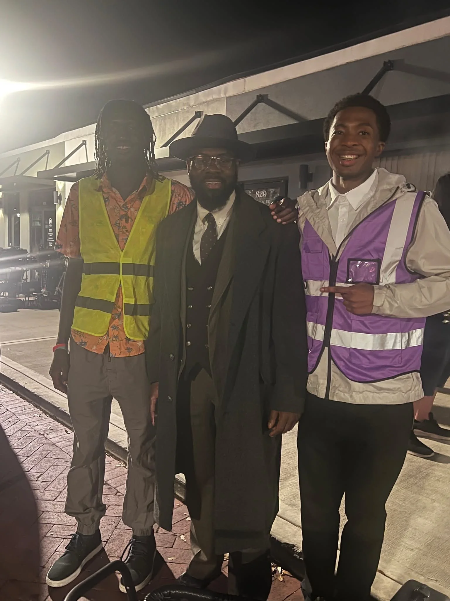 Three men standing together outdoors at night, smiling for the camera. One is wearing a colorful shirt and yellow safety vest. Another is in a black suit with a hat and glasses. The third man is wearing a white shirt with a purple and white jacket.