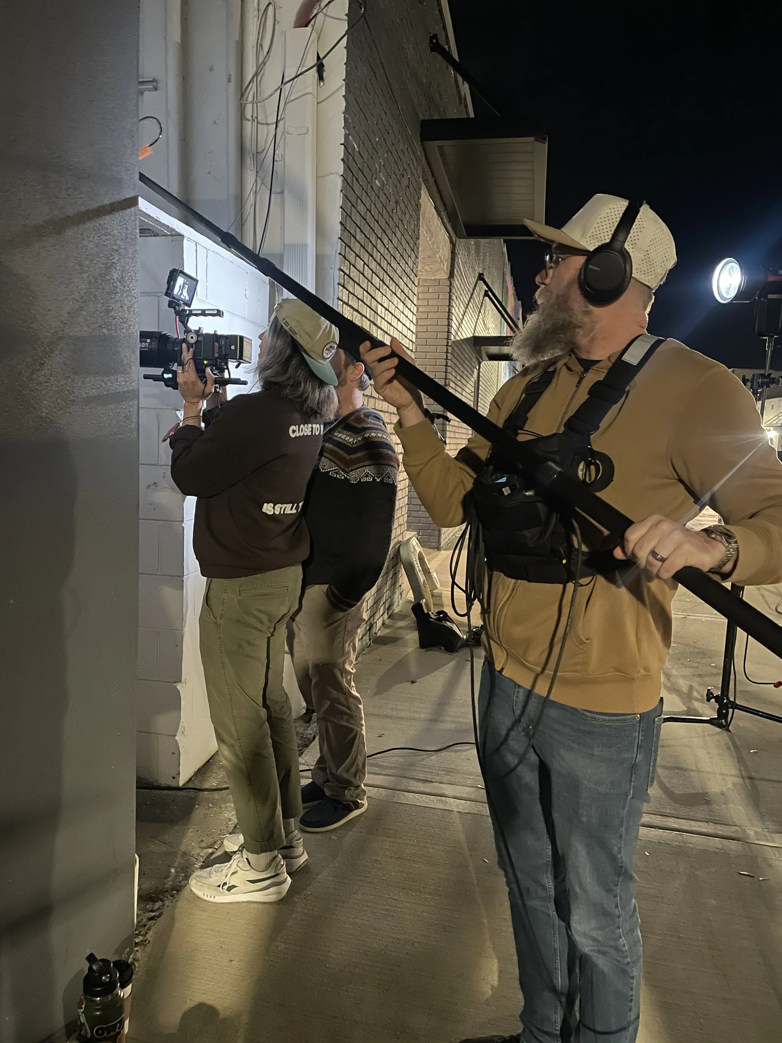 People engaged in filming or photography during nighttime outside a building in Clermont with brick and concrete walls, some holding cameras and gear, with one man wearing headphones and a beige cap.
