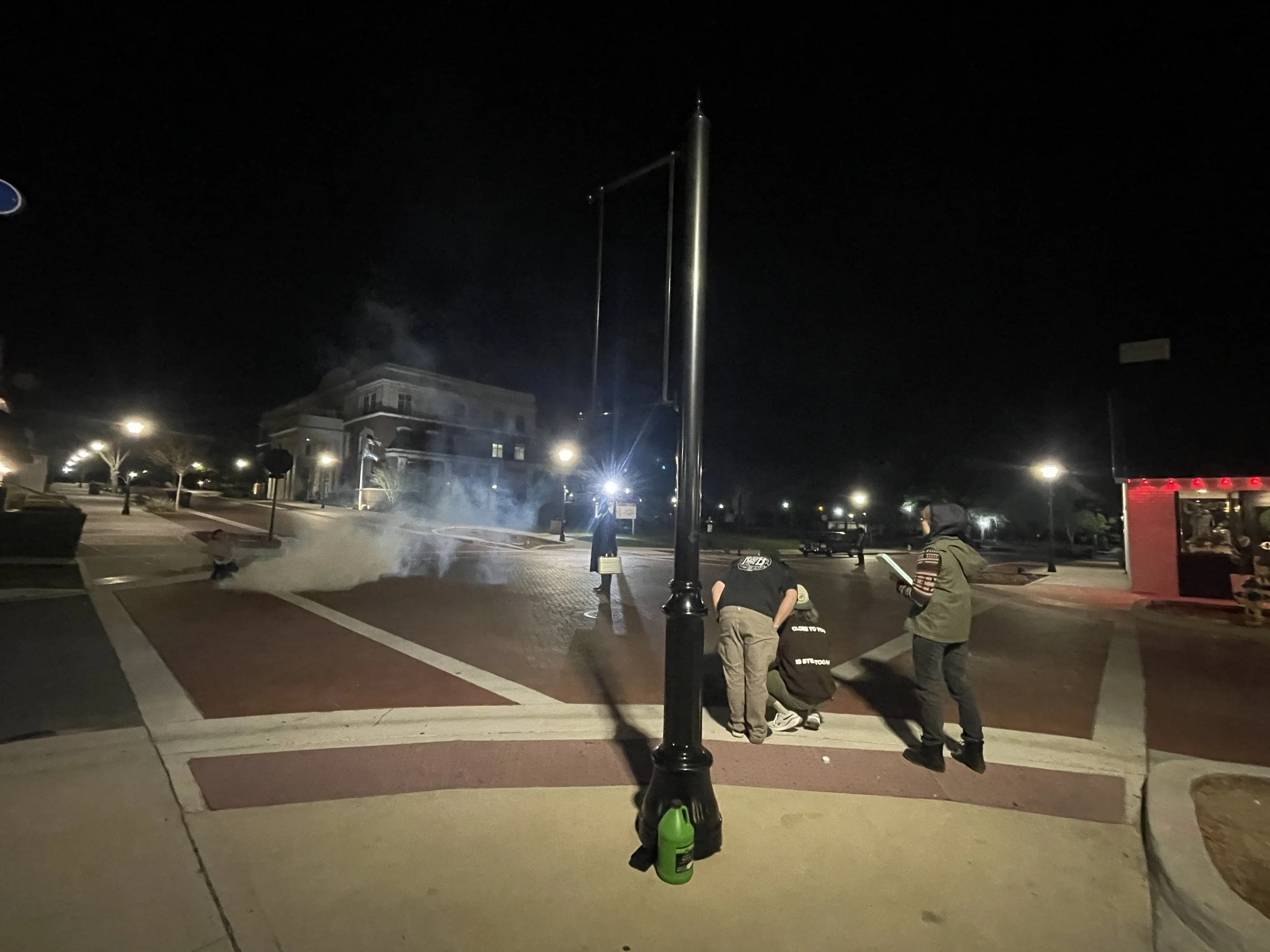 Nighttime scene in a town square in Clermont with a flagpole, a few people crouching on the ground, and a building with lit windows in the background.