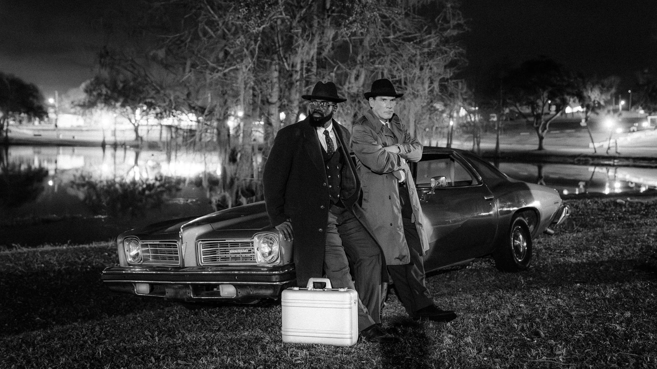 Two men in dark clothing wearing hats, standing by a vintage car at night in a park, with a river, trees, and distant lights in the background.