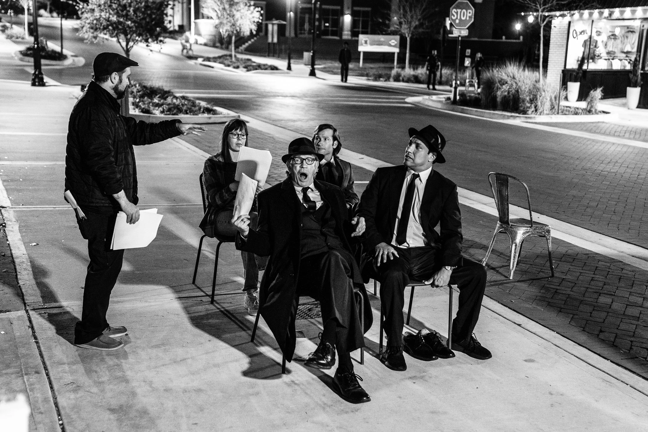 A group of five people, four men and one woman, sitting on chairs at a sidewalk corner at night, with one man standing and talking. The group appears to be engaged in a discussion. The scene is in black and white, with streetlights illuminating the a