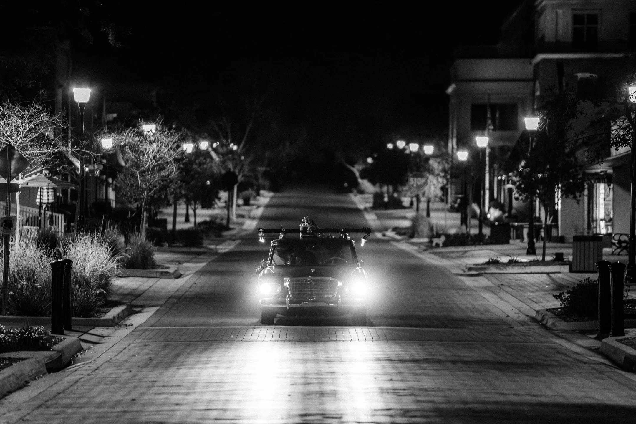 A black and white photo of a deserted street at night with a vintage car approaching from the distance with headlights on. The street is lined with trees, lampposts, and residential buildings, with no visible people.