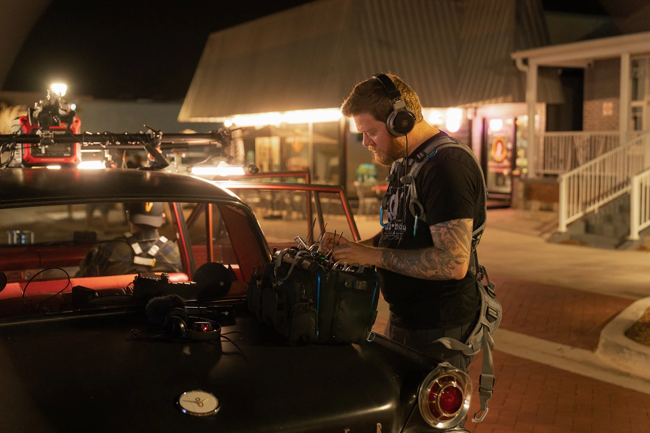 A man with a beard and tattoos on his arms working on electronic equipment at night outside a building with warm lighting, a porch, and a vending machine in the background.