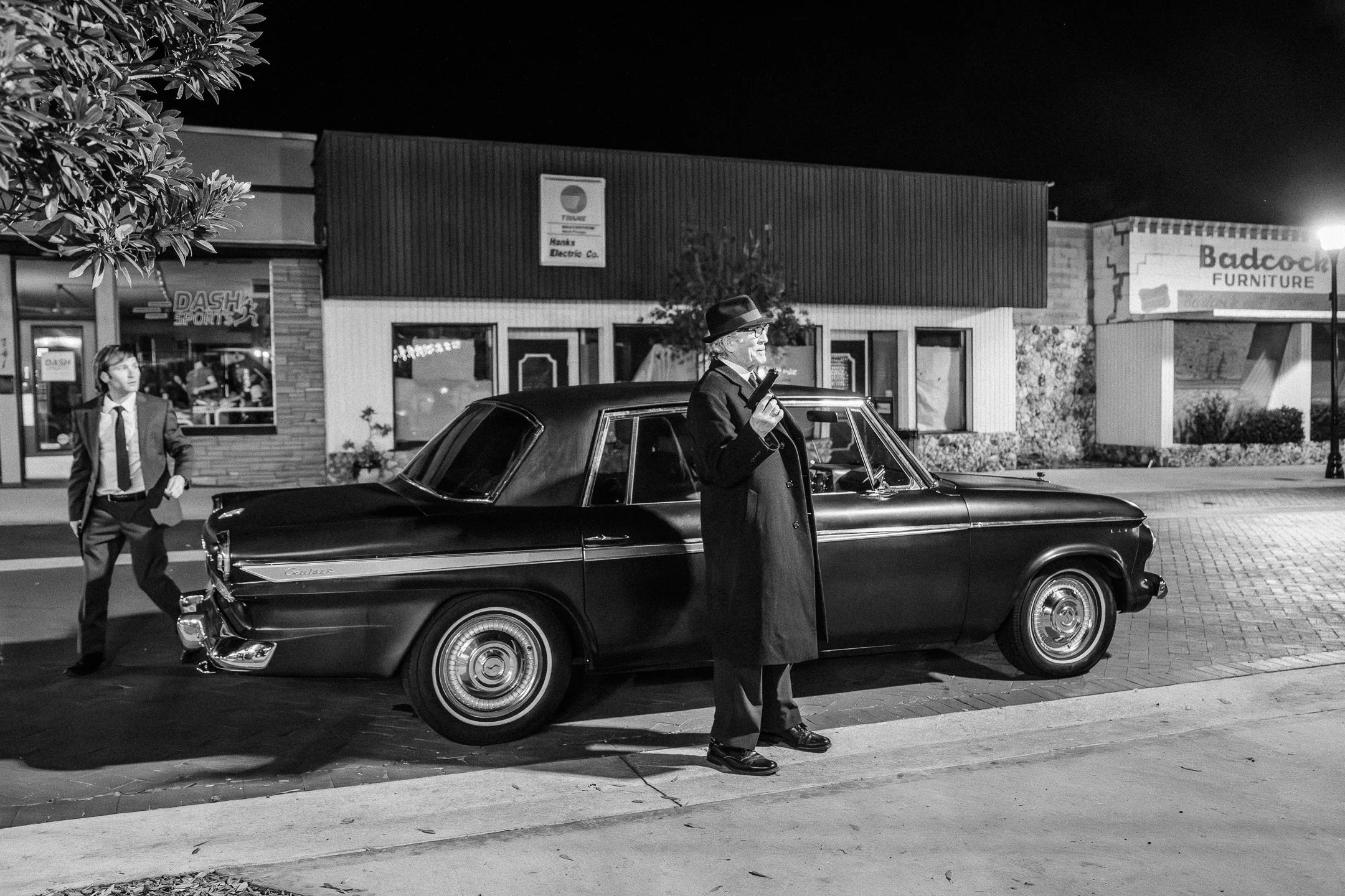 A black-and-white photo of a man in a suit and fedora hat standing next to a vintage black car at night. A young man in a suit is walking nearby. In the background, there are shops with lit signs, including one for Badcock Furniture.