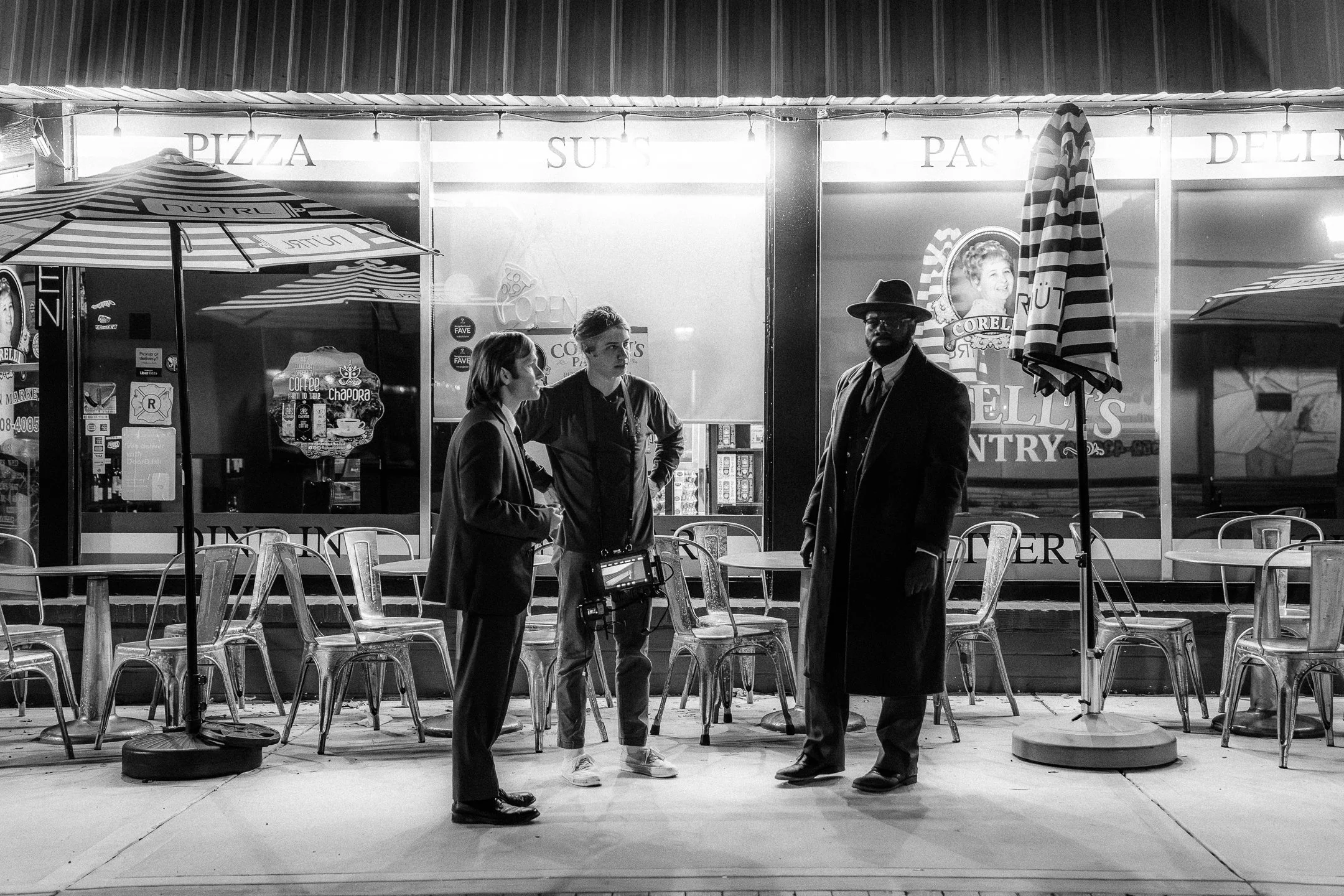Three people standing and talking in front of a restaurant with outdoor seating, umbrellas, and signs for pizza, sushi, pasta, and gelato, at night.
