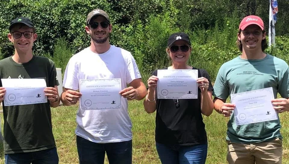 Four people standing outdoors on grass, each holding a certificate and smiling. They are dressed casually and are wearing sunglasses and caps. There are green trees in the background.