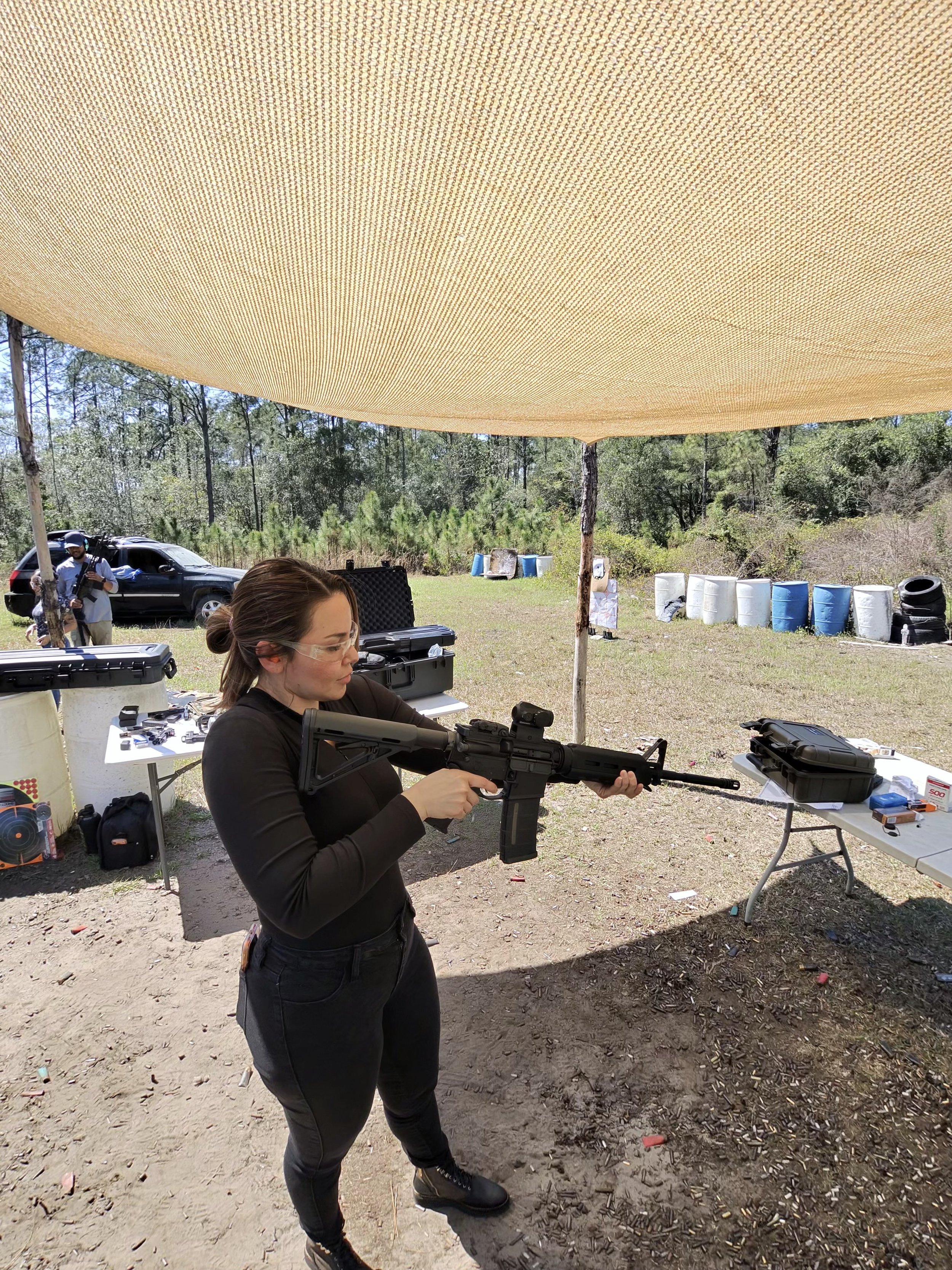 A woman in black clothing and sunglasses holding a rifle at an outdoor shooting range, with targets and containers in the background.
