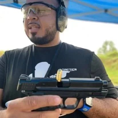 Man wearing safety glasses, hearing protection, and black T-shirt holding a handgun at an outdoor shooting range.
