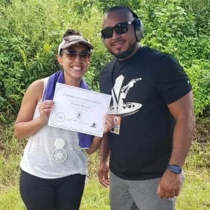 A woman holding a certificate and a man standing beside her outdoors on a sunny day, both smiling.