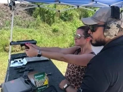 A woman aiming a handgun under supervision at an outdoor shooting range, with a man assisting her, both wearing protective eyewear and a shaded tent overhead.