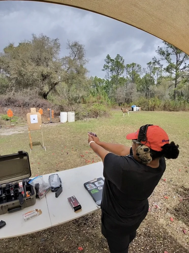 A person at an outdoor shooting range aiming a handgun at targets downrange. They are wearing earmuffs, a cap, and protective glasses, with a table holding firearm accessories nearby.