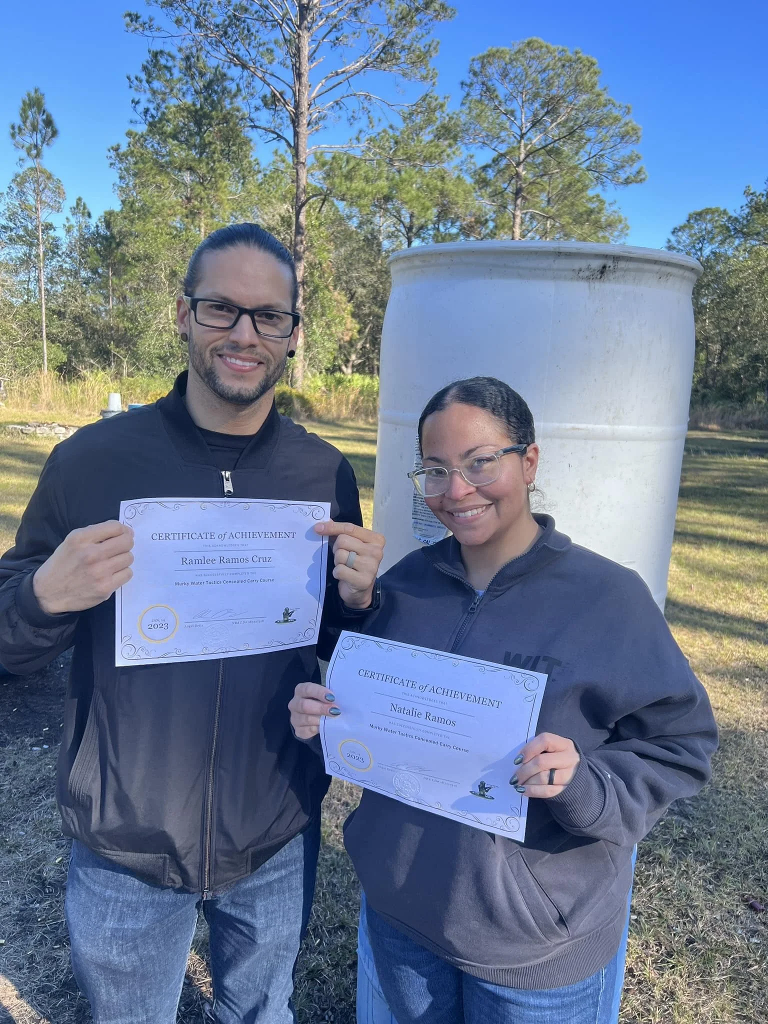 Two women outdoors holding certificates of achievement, smiling, with trees and a blue sky in the background.
