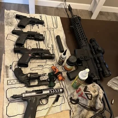 A table with three handguns, a rifle with a scope, various cleaning supplies, a cloth, and small accessories.