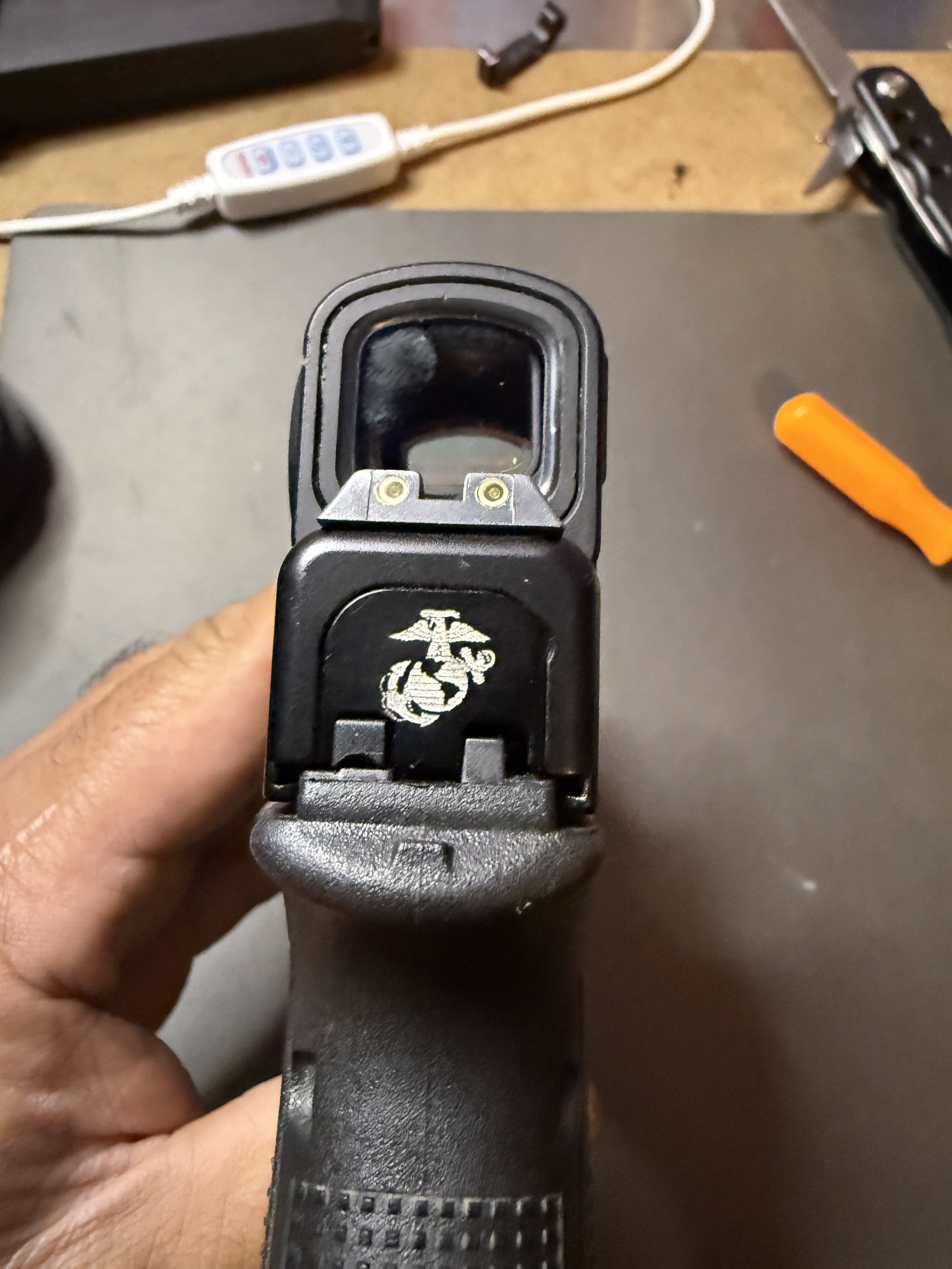 Close-up of a person's hand holding a black firearm, viewed from above, with a black slide and rubber grip. The background includes orange and black tools, a white power strip, and a tan surface.
