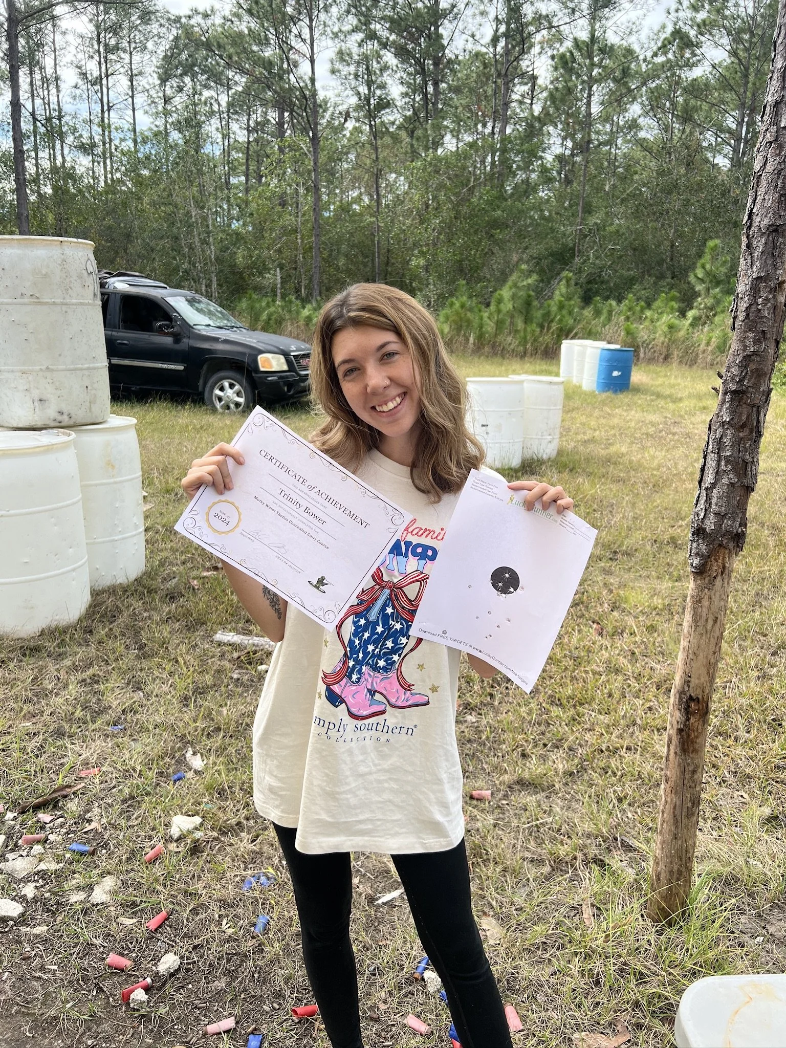 A young woman smiling while holding certificates outdoors with trees, a black vehicle, and scattered shotgun shells on the ground.