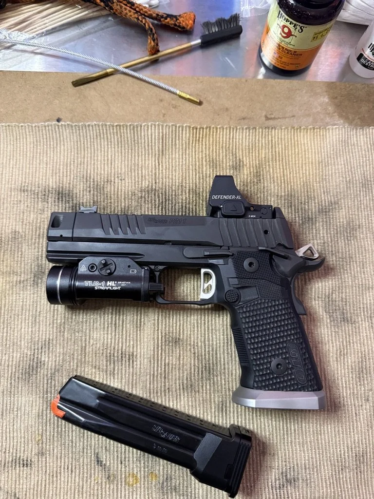 A black semi-automatic handgun with a flashlight attached to the rail, a magazine, and a mounted red dot sight. The objects are placed on a tan cloth, with cleaning brushes, a bottle of gun cleaning solvent, and cleaning tools visible in the background.