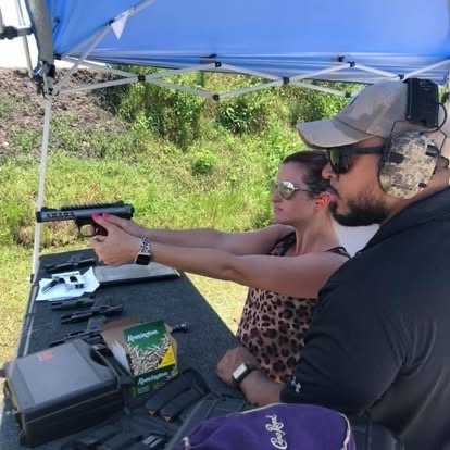 Woman holding and aiming a handgun while sitting at a table with a man at an outdoor shooting range, under a blue canopy.