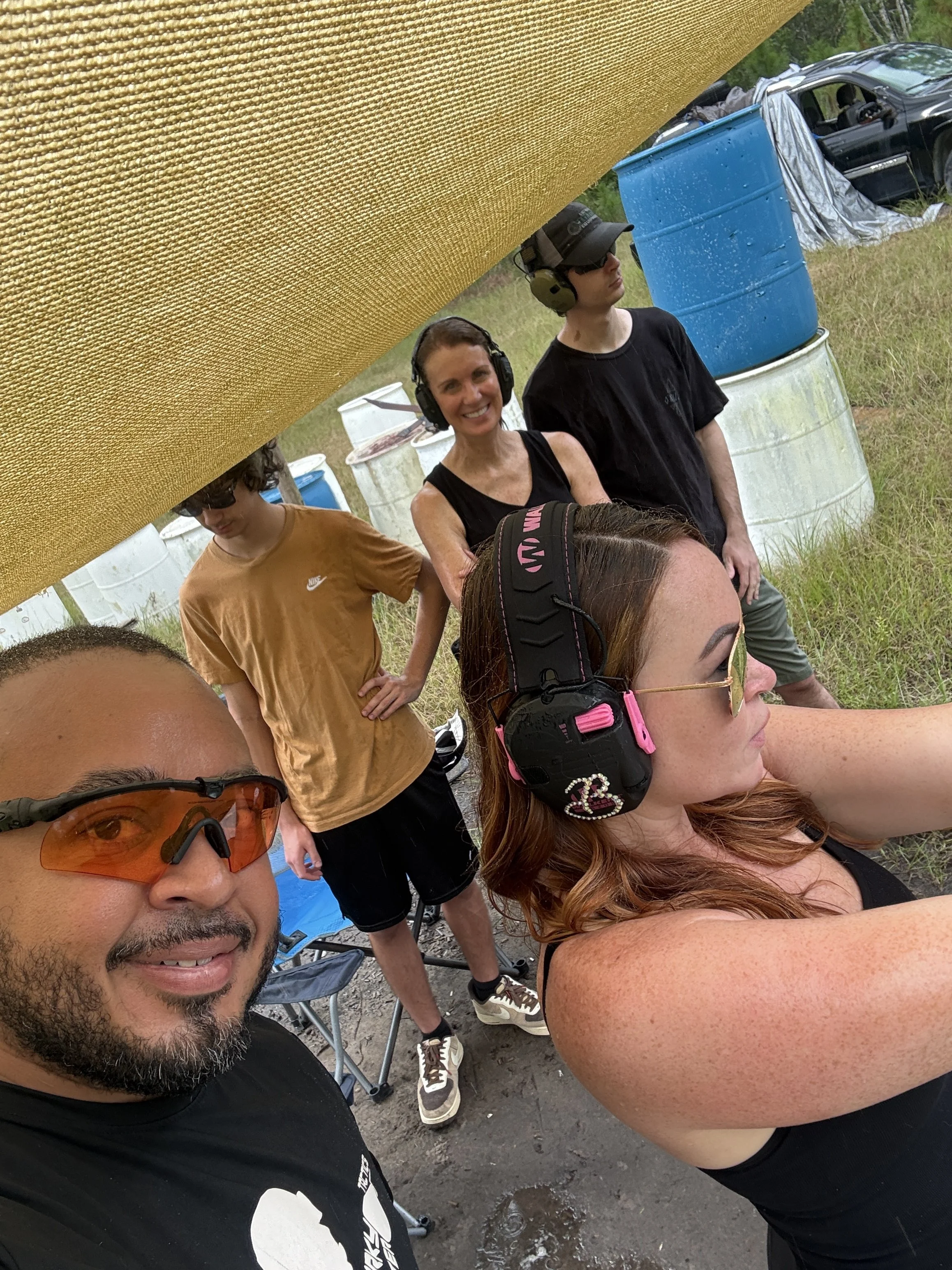 Group of people taking a selfie outdoors, with a large yellow shade cloth above them and barrels in the background.