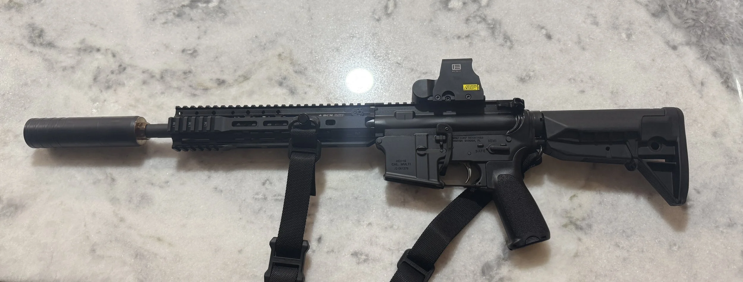 A black tactical rifle with a silencer, red dot sight, adjustable stock, and sling, laid on a light-colored surface.