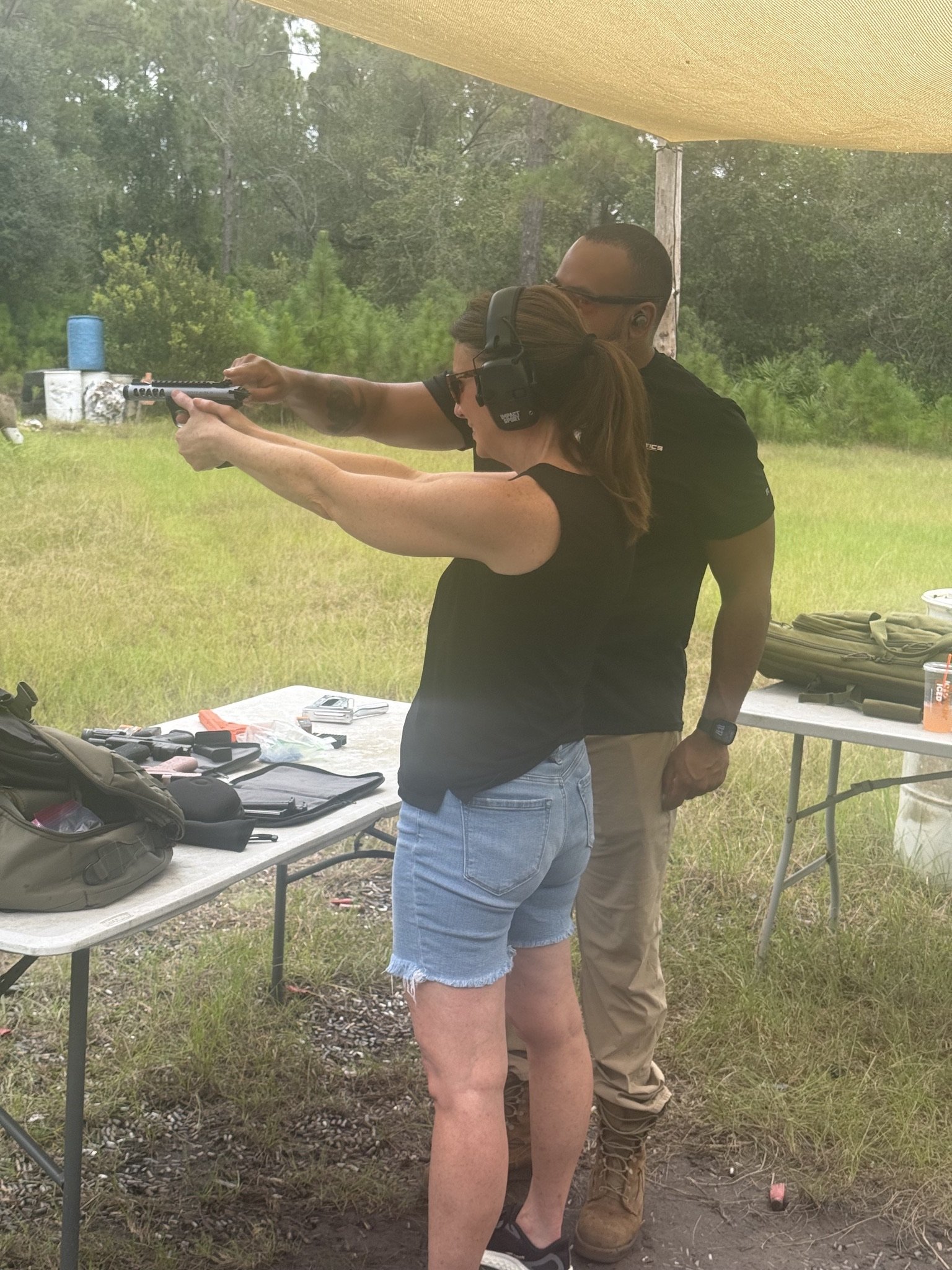A woman shooting a handgun with the assistance of a man at an outdoor shooting range, wearing ear protection and sunglasses, with tactical gear and a table with equipment nearby.