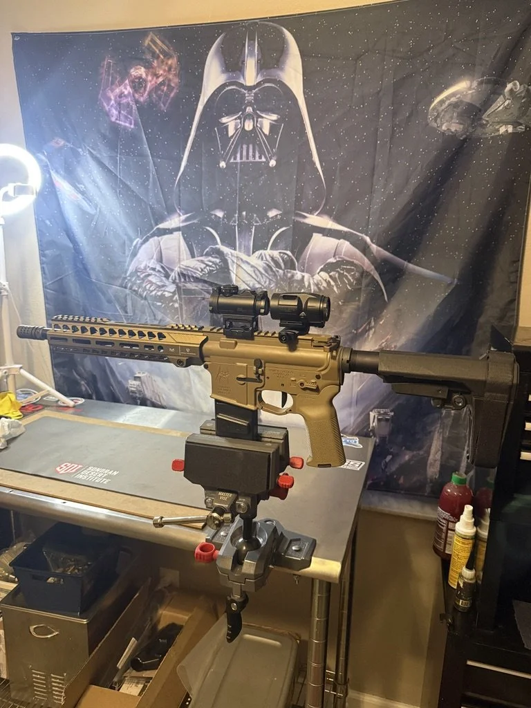 A custom rifle with a scope mounted on a rest, set up on a workbench in front of a large Darth Vader poster with a Star Wars theme. The workbench has tools and supplies on it.