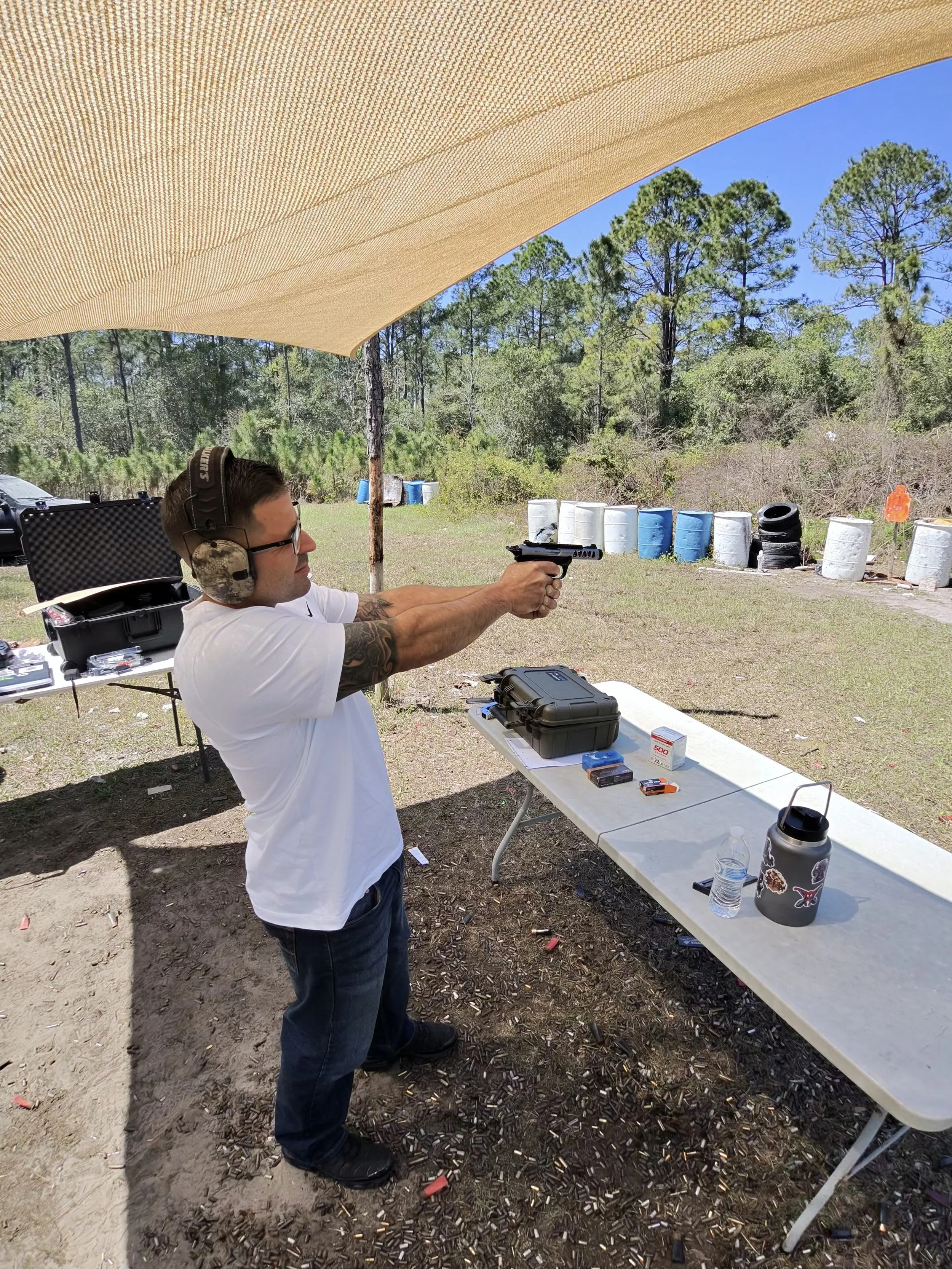 A man at an outdoor shooting range is aiming a handgun while wearing ear protection and glasses. There are boxes of ammunition on a nearby table, and spent cartridge casings scattered on the ground. Several barrels and trees are visible in the backgr