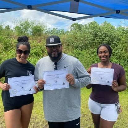 Three people standing outdoors under a blue canopy, holding certificates and smiling. Two women on each side of a man, all dressed casually, with green trees in the background.