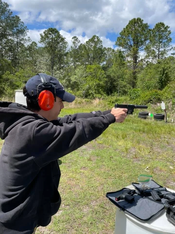 A person wearing a black hoodie, navy cap, and red hearing protection, aiming a pistol at an outdoor shooting range with trees and cloudy sky in the background.