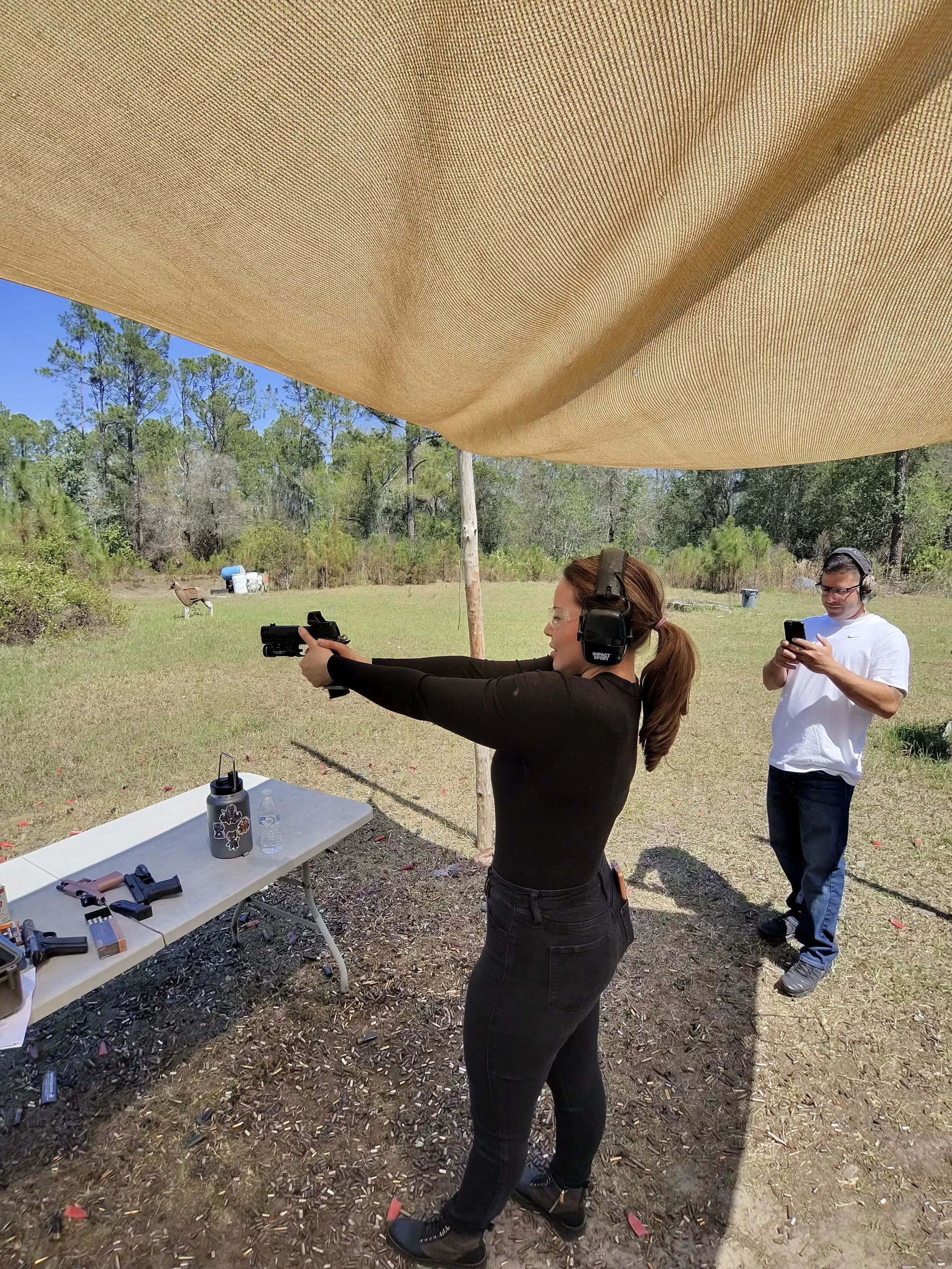 A woman with brown hair in a ponytail, wearing black clothing, aiming a gun with both hands under a large beige canopy outdoors. A man in a white shirt and glasses is standing nearby, looking at his phone. A table with various firearms and items is i