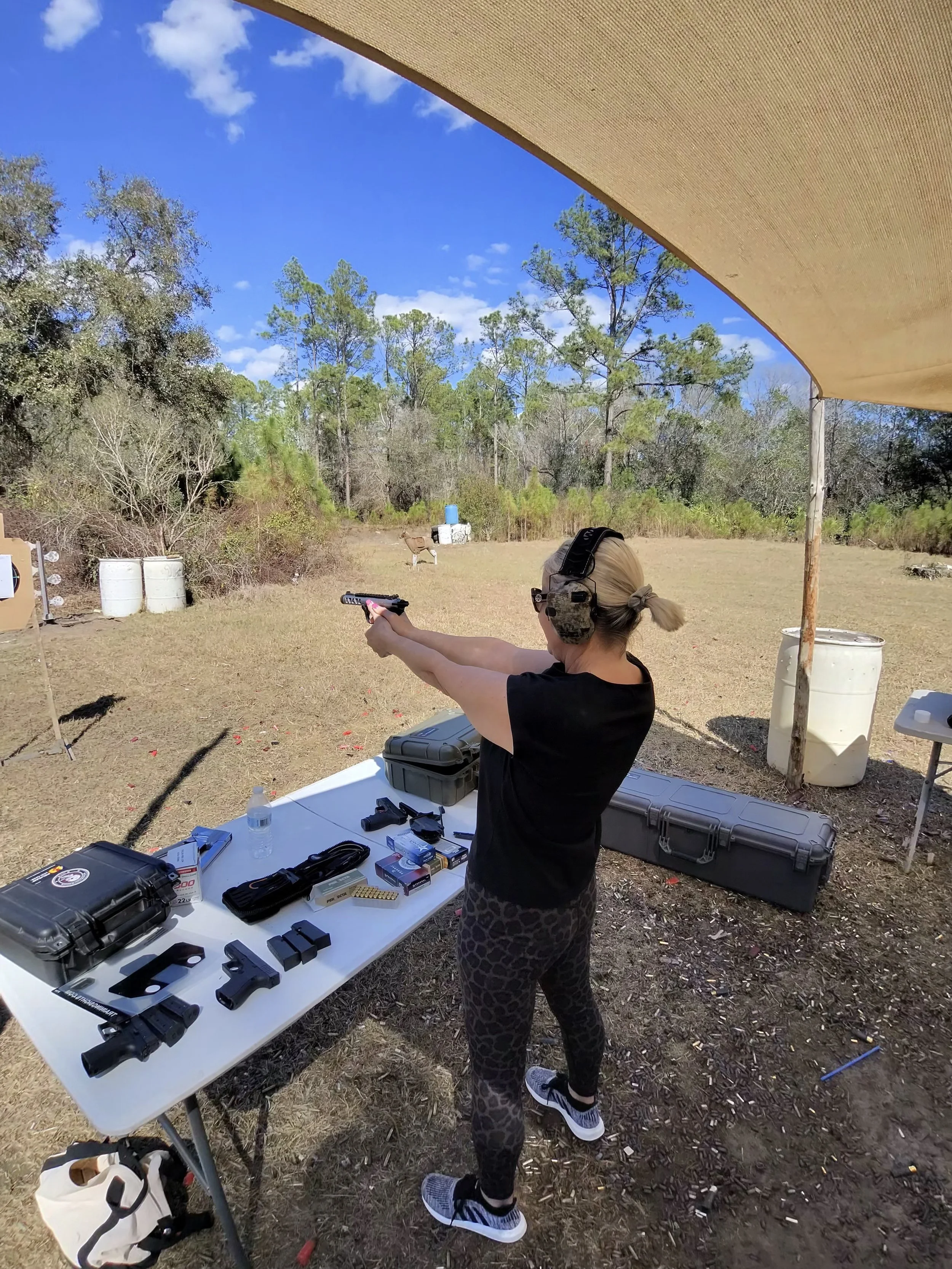 A woman at an outdoor shooting range aiming a handgun, wearing ear protection, standing behind a table with firearms, ammunition, and shooting accessories, under a canopy with trees and blue sky in the background.