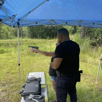 A man holding a gun pointed at a woman under a blue canopy tent outdoors in a grassy area with trees in the background.
