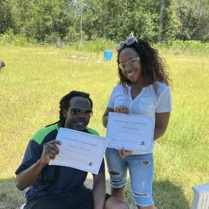 Two people outdoors, one sitting and holding a certificate, the other standing and also holding a certificate, in a grassy field with trees in the background.