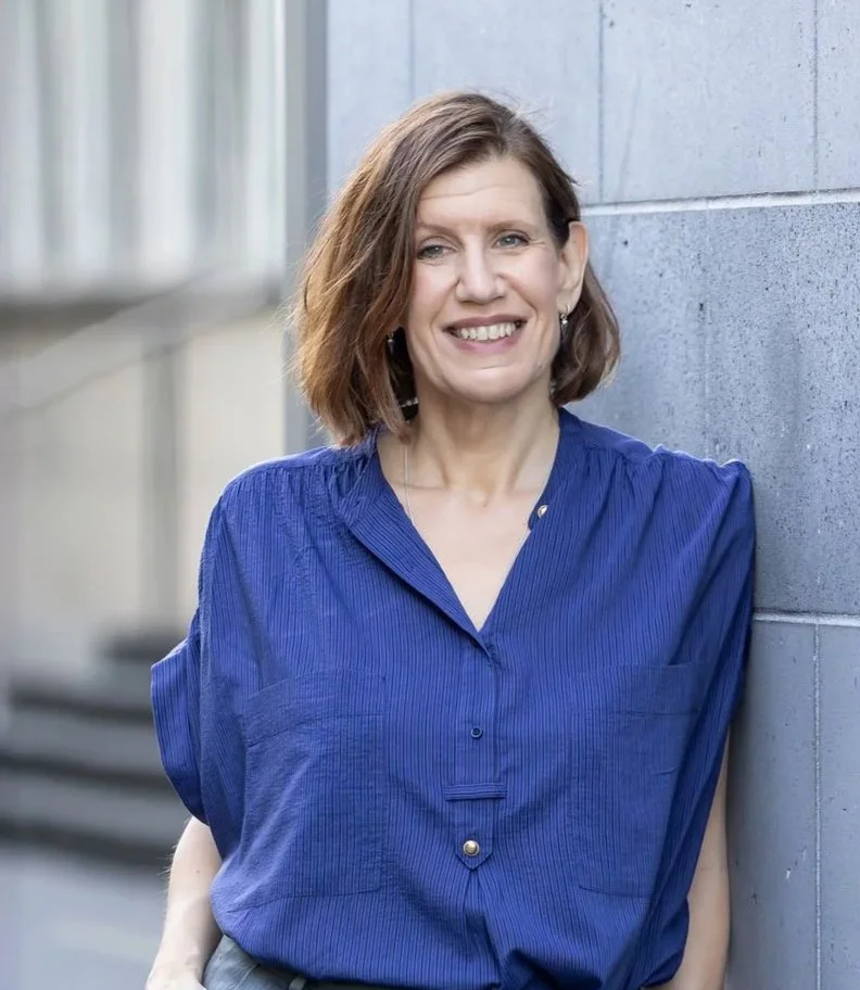 A woman with shoulder-length brown hair, wearing a blue blouse, smiling and leaning against a gray concrete wall outdoors.