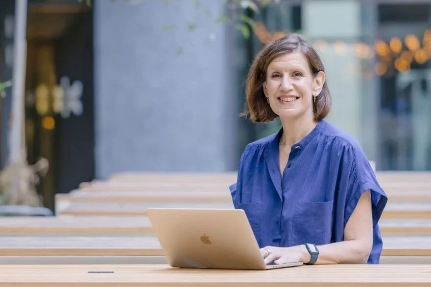 A woman in a blue blouse sitting at an outdoor table with a silver laptop, smiling at the camera.