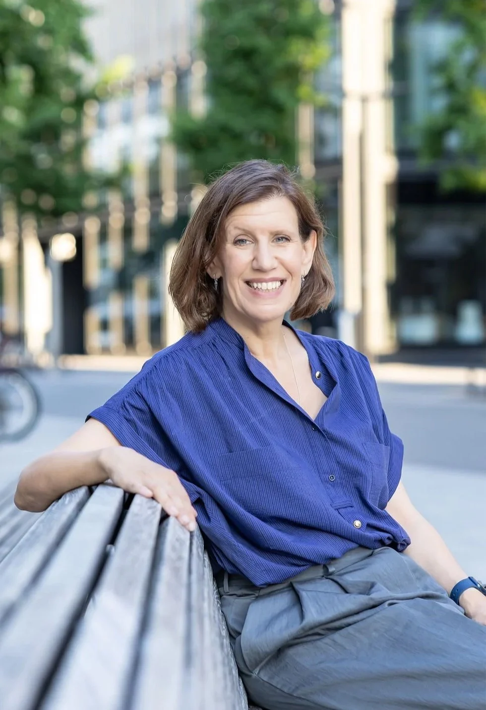 A woman sitting on a bench outdoors, smiling, wearing a blue shirt and gray pants, with green trees and buildings in the background.
