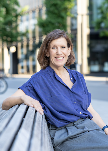 A smiling woman with shoulder-length brown hair, wearing a blue button-down shirt and gray pants, sitting on a park bench in downtown Zurich  with trees and buildings in the background.