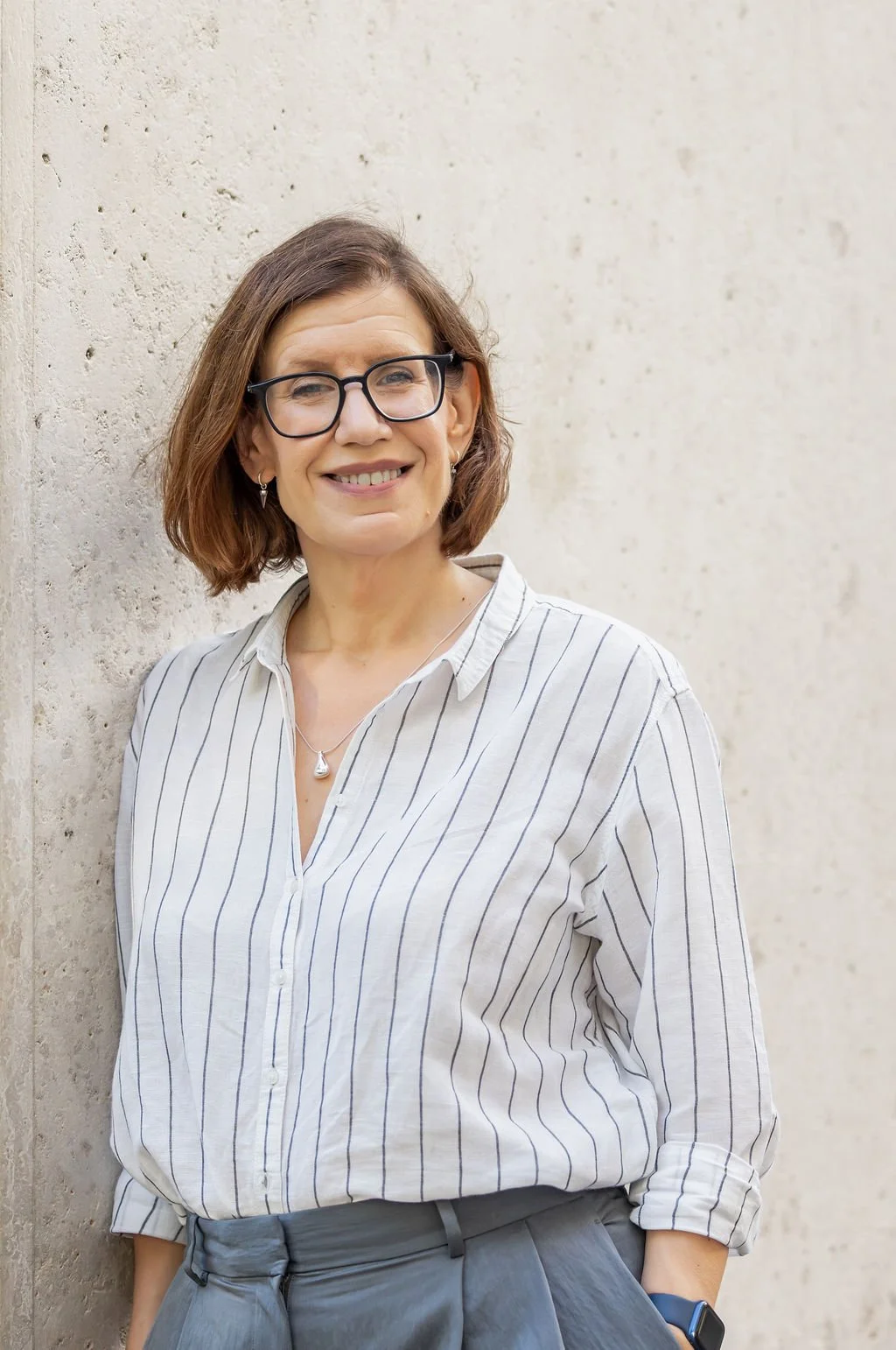 A woman with shoulder-length brown hair, wearing glasses, a striped white shirt, and gray pants, standing against a beige textured wall with a slight smile.