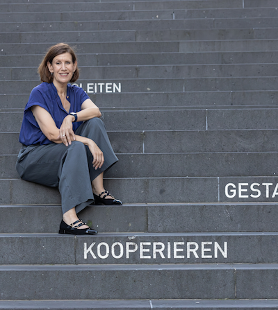 A smiling woman with short brown hair sitting on concrete steps with German words 'Reiten,' 'Geste,' and 'Kooperieren' written on the steps.