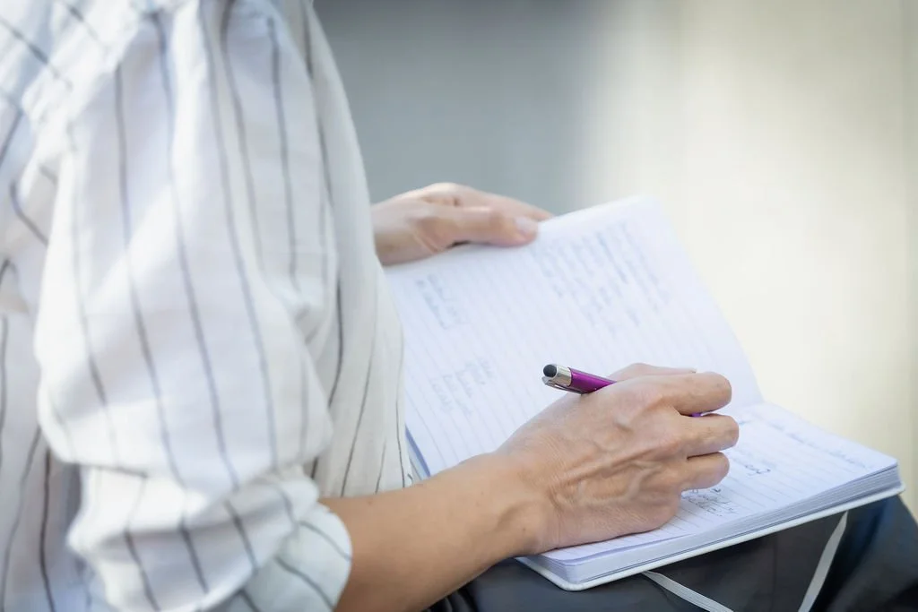 A person wearing a striped shirt is writing in a notebook with a purple pen.