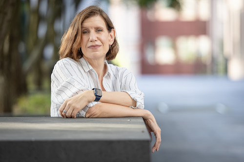 A woman with short brown hair and a striped shirt, leaning on a concrete surface outdoors, looking contemplative.