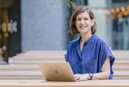 A woman sitting at a wooden table outdoors with a laptop, smiling at the camera.