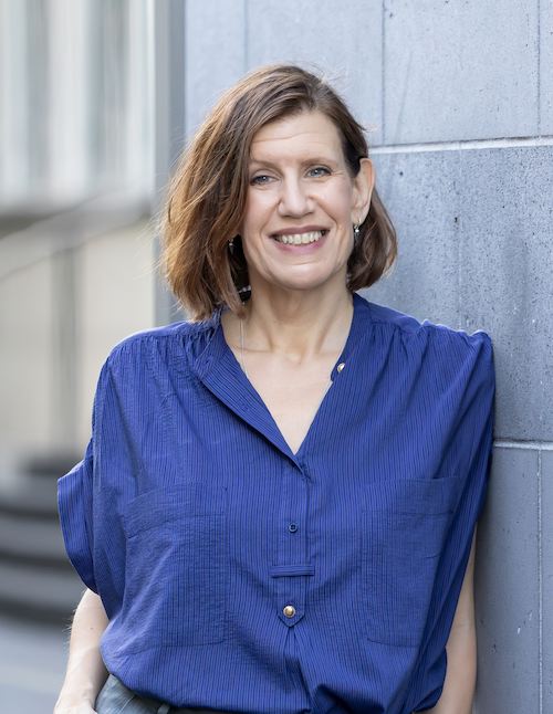 Woman with brown hair dressed in bloue shirt leaning against wall in Zurich