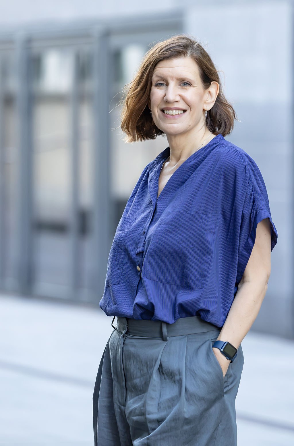 A woman with shoulder-length brown hair, wearing a blue blouse and gray pants, standing outside near a modern building, smiling at the camera.