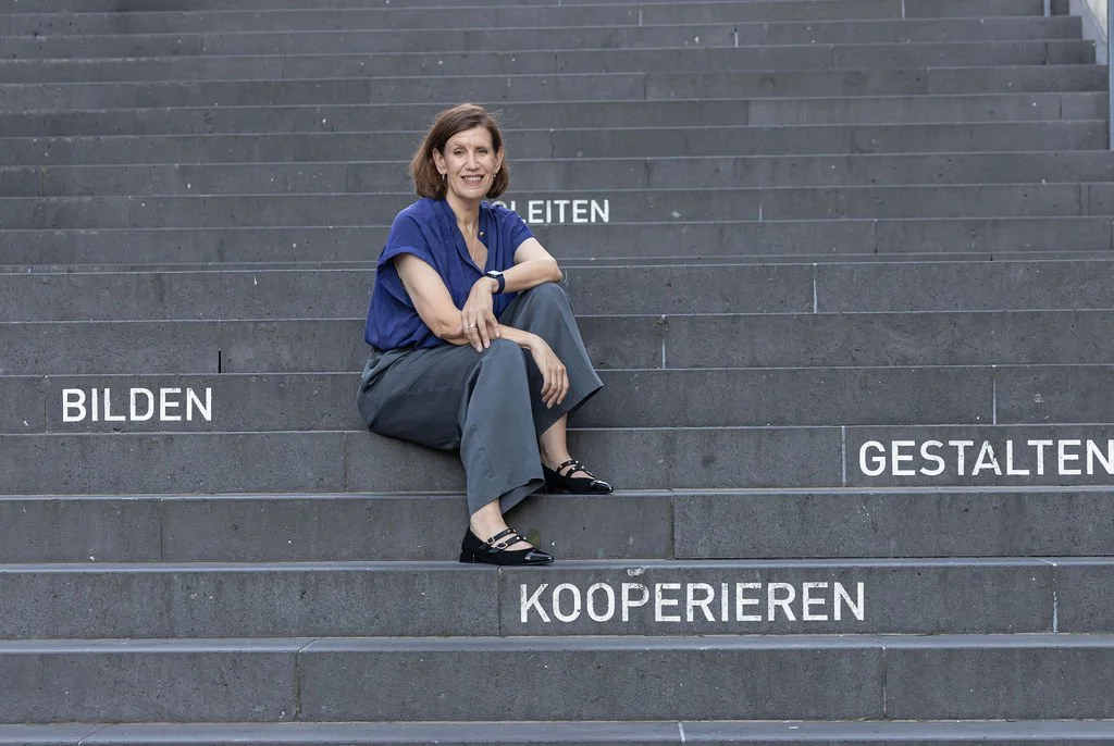 Woman sitting on stairs with German words painted on steps, including 'BILDEN,' 'GLEITEN,' 'GESTALTEN,' and 'KOOPIEREN'.