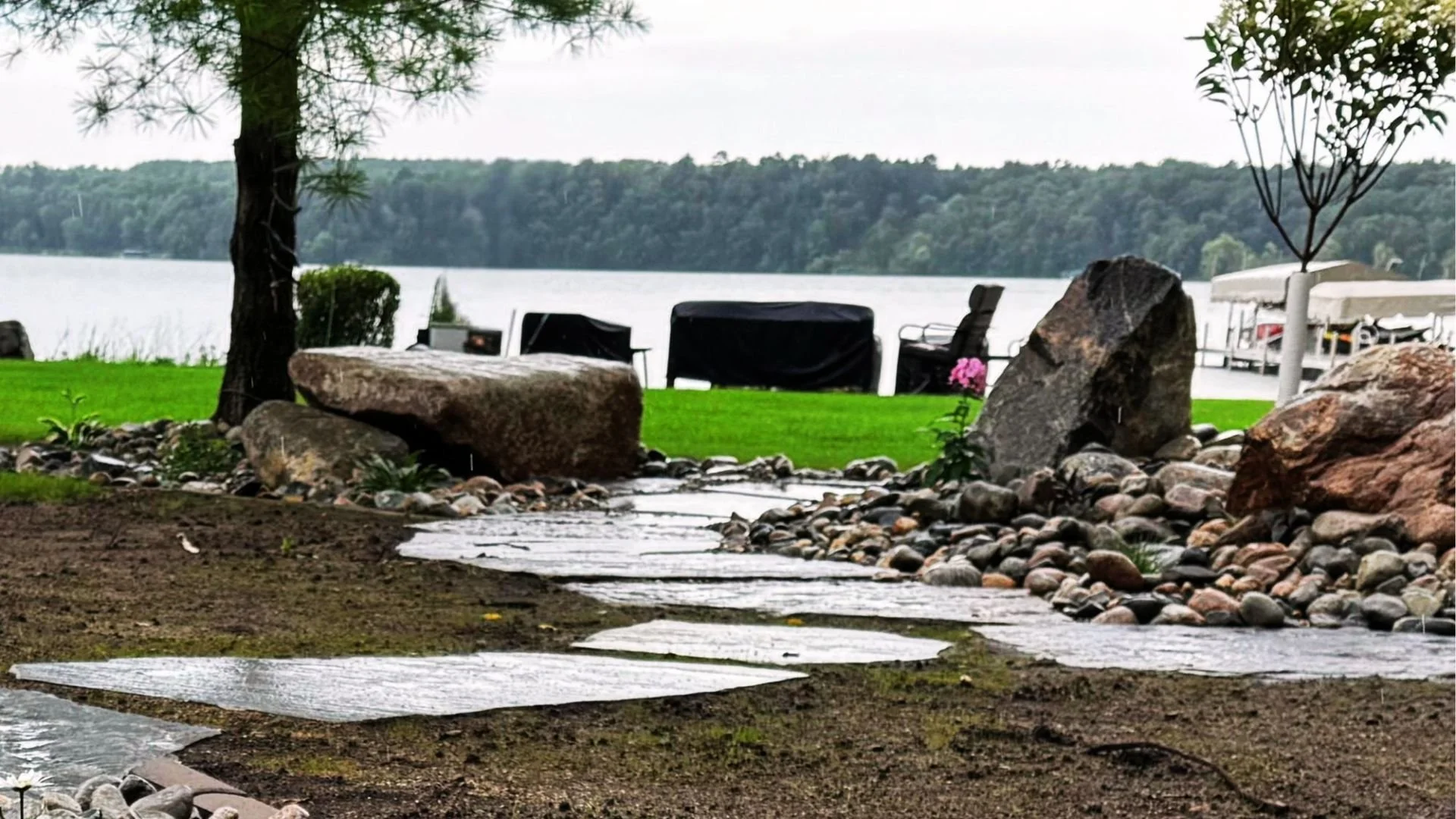 A lakeside yard scene with large rocks, a small stream, green grass, trees, lounge chairs, and a boat dock in the background.