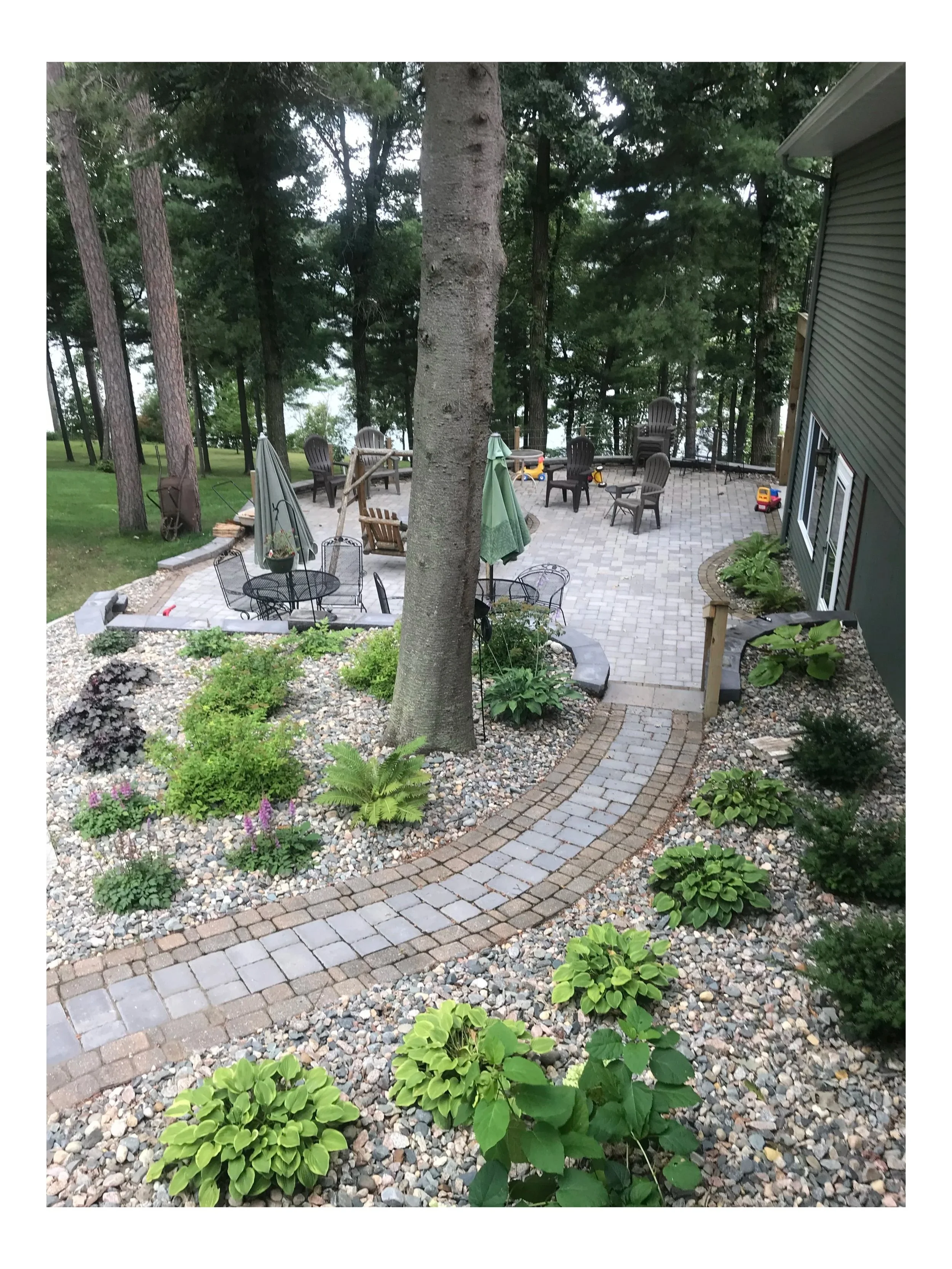 A backyard patio area with a stone pathway, surrounded by small green plants and flowers, with a large tree in the center and outdoor furniture including chairs, a table, and umbrellas, outdoors next to a house with green siding.